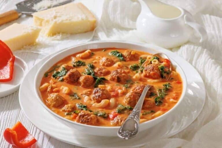 A bowl of meatball soup with pasta, spinach, and tomato broth, served with a spoon.