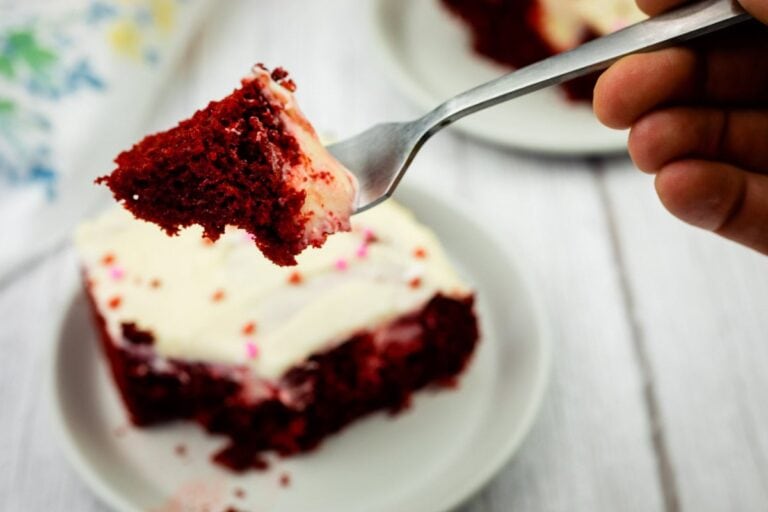 A hand holds a fork with a bite of red velvet cake topped with white frosting above a plate with the remaining cake.