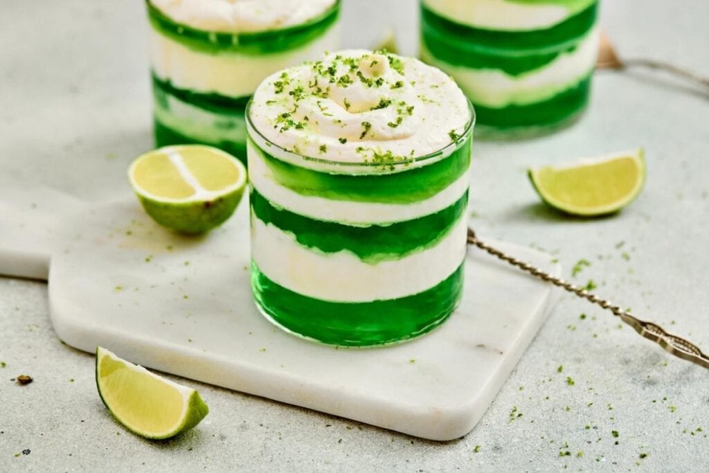 A glass dessert of St. Patrick’s Day Green Jello Parfaits, sits on a marble board with lime wedges nearby.