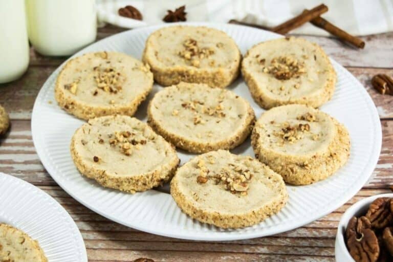 A white plate holds eight round pecan cookies, each topped with chopped pecans, on a wooden surface with milk bottles and cinnamon sticks in the background.