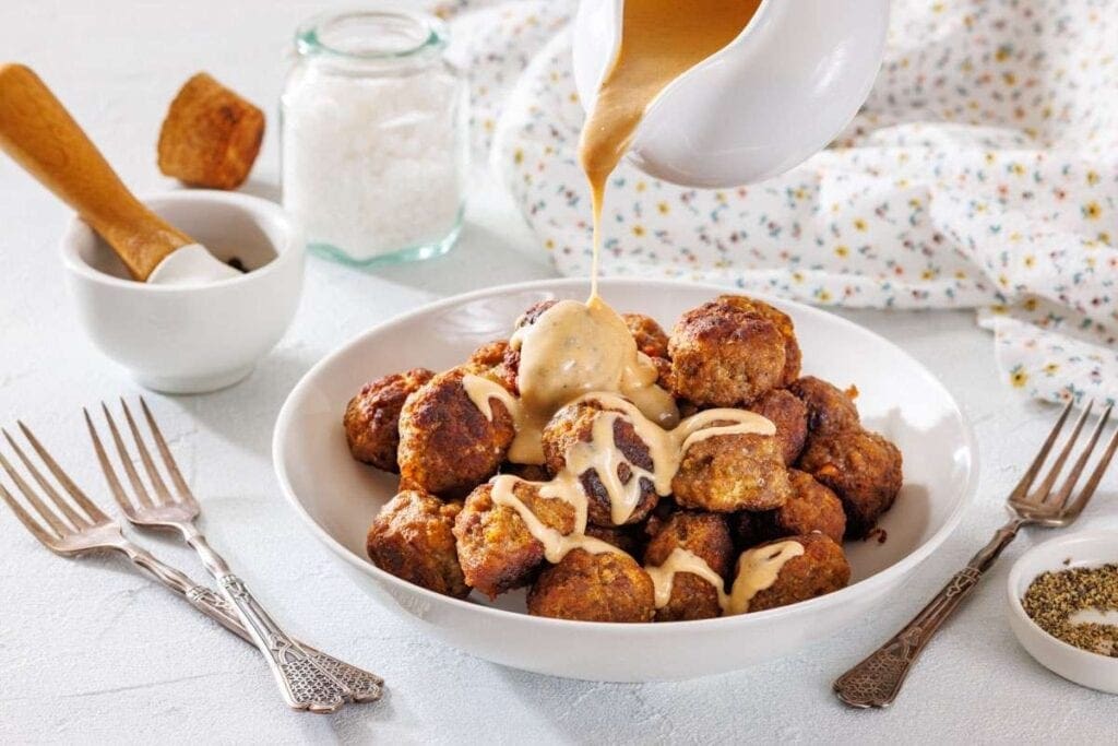A bowl of meatballs is topped with creamy sauce being poured from a jug, surrounded by forks and seasoning jars on a white table.