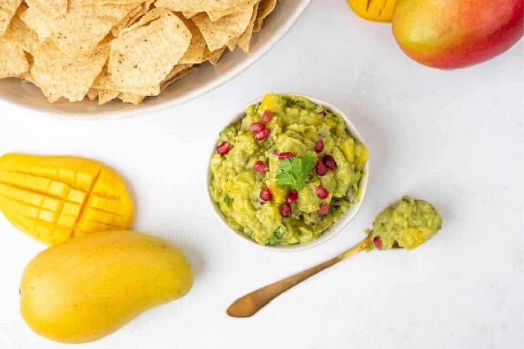 A bowl of Tropical mango guacamole sits next to a spoon, tortilla chips, and two mangoes on a white surface.