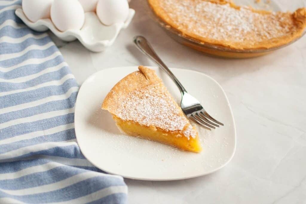 A slice of vinegar pie dusted with powdered sugar on a white plate with a fork, next to a blue striped cloth and a dish of eggs.