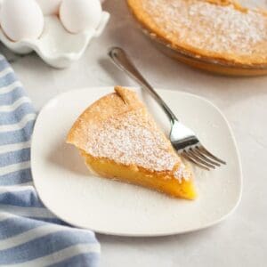 A slice of vinegar pie dusted with powdered sugar on a white plate with a fork, next to a blue striped cloth and a dish of eggs.