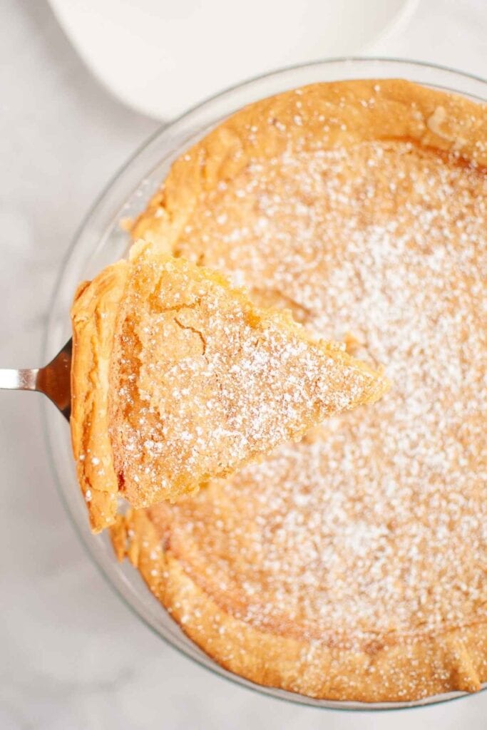 A slice of golden brown pie dusted with powdered sugar is being lifted from a whole pie on a glass plate.