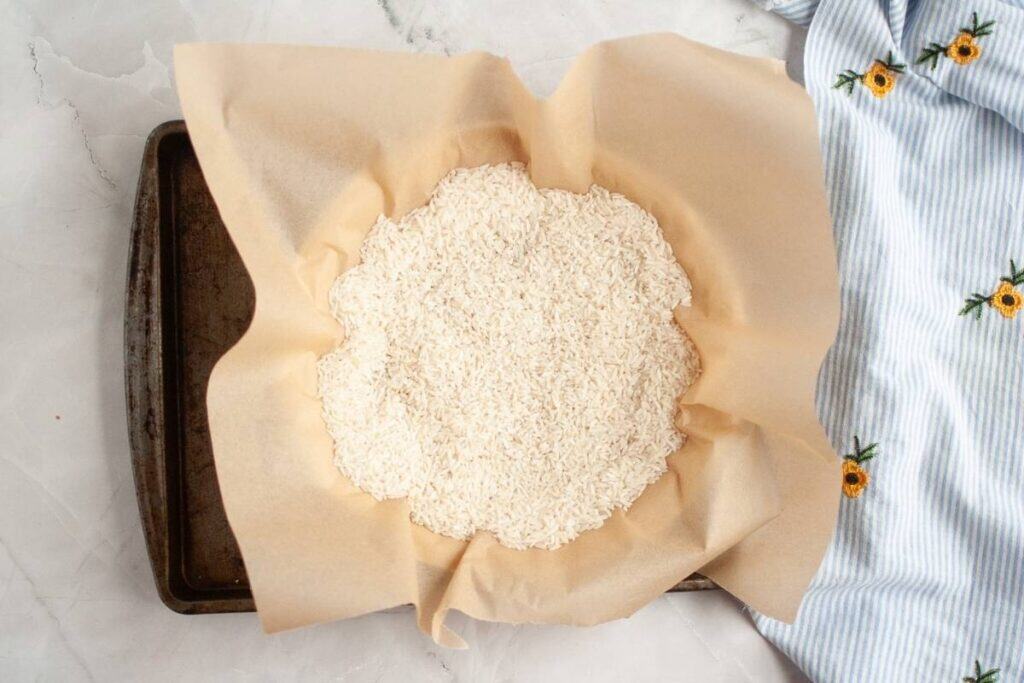 A round cake pan lined with parchment paper filled with uncooked white rice, placed on a metal baking sheet next to a blue fabric with yellow flower embroidery.