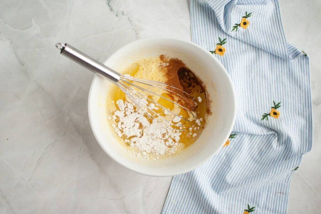 A mixing bowl with a whisk, containing eggs, flour, and spices next to a blue and white striped cloth with yellow flower embroidery.