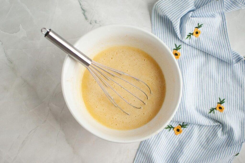 A metal whisk rests in a white bowl filled with a light batter, next to a blue-and-white striped cloth embroidered with yellow flowers.