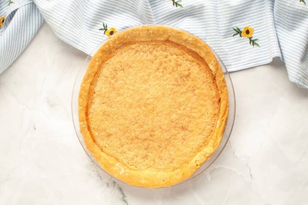 A baked pie with a golden, slightly textured top sits on a white surface next to a blue and white striped cloth with yellow embroidered flowers.