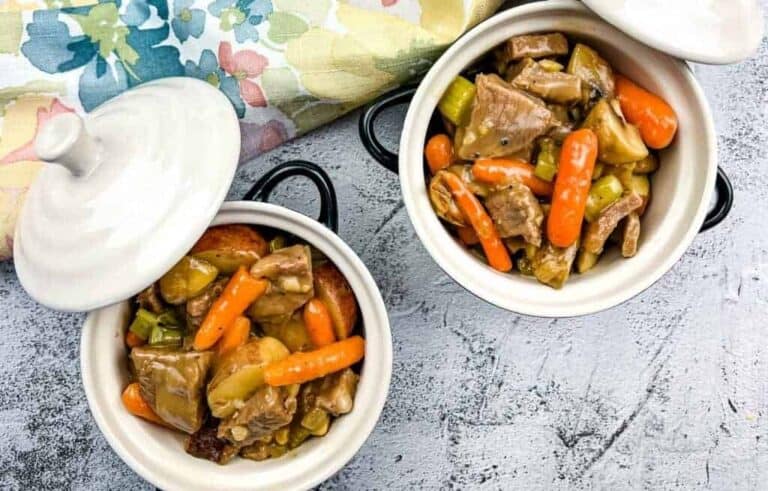 Two white ceramic bowls filled with beef stew containing pieces of meat, baby carrots, celery, and potatoes, placed on a gray surface next to a floral napkin.