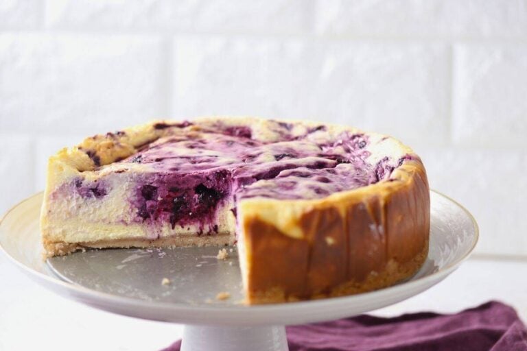 A round blueberry swirl cheesecake with a slice removed, displayed on a white cake stand against a white background.