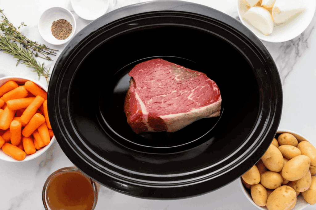 Raw beef roast in a slow cooker surrounded by bowls of carrots, potatoes, onion, broth, herbs, and seasonings on a white countertop.