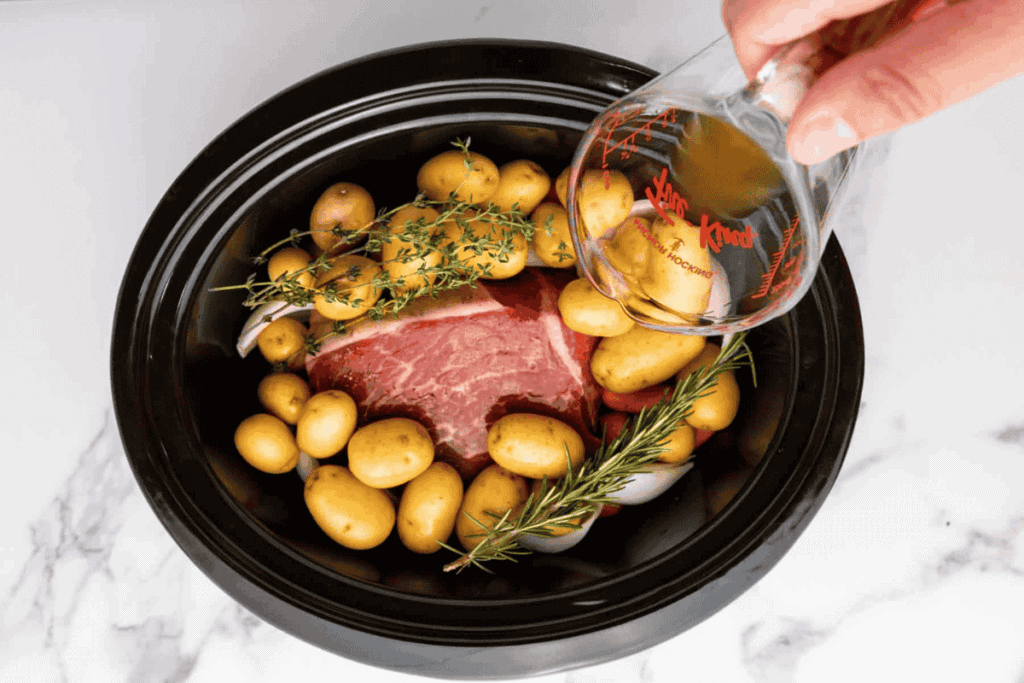 A hand pours liquid from a measuring cup into a slow cooker containing a raw meat cut, baby potatoes, onions, rosemary, and thyme.