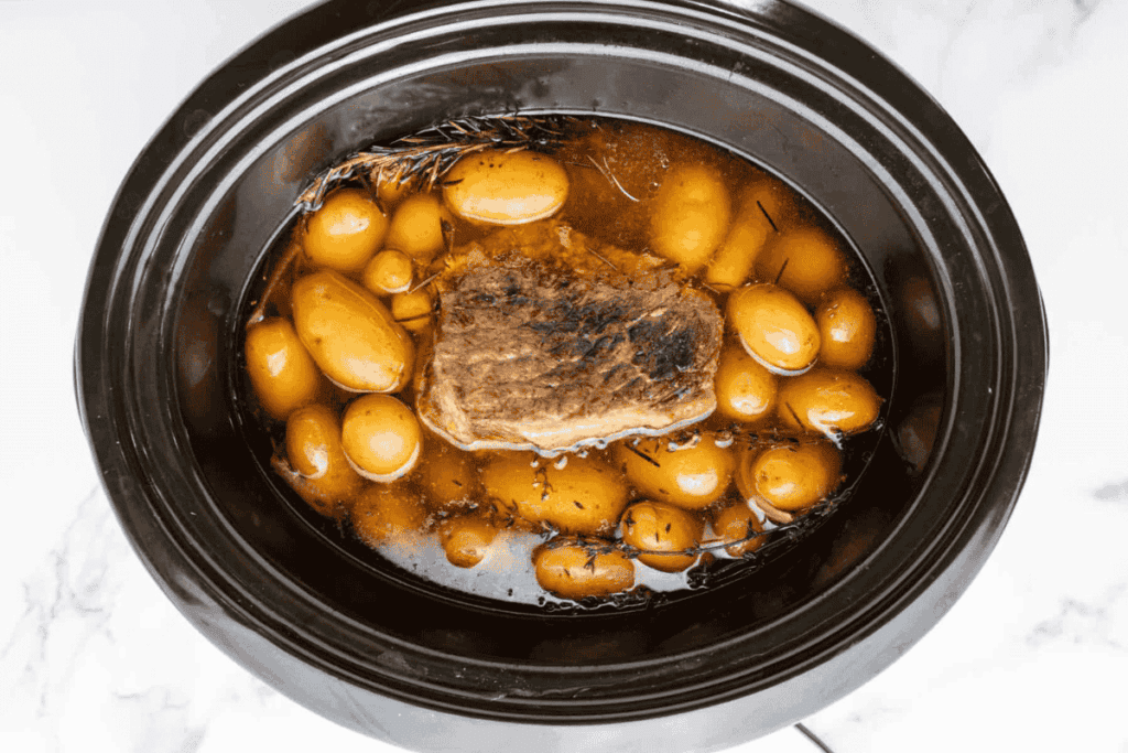 A slow cooker filled with a cooked beef roast, whole baby potatoes, and broth, with sprigs of herbs floating in the liquid.