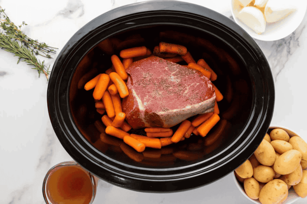 A raw beef roast sits on top of baby carrots in a slow cooker, surrounded by bowls of potatoes, garlic cloves, rosemary, and broth on a marble countertop.