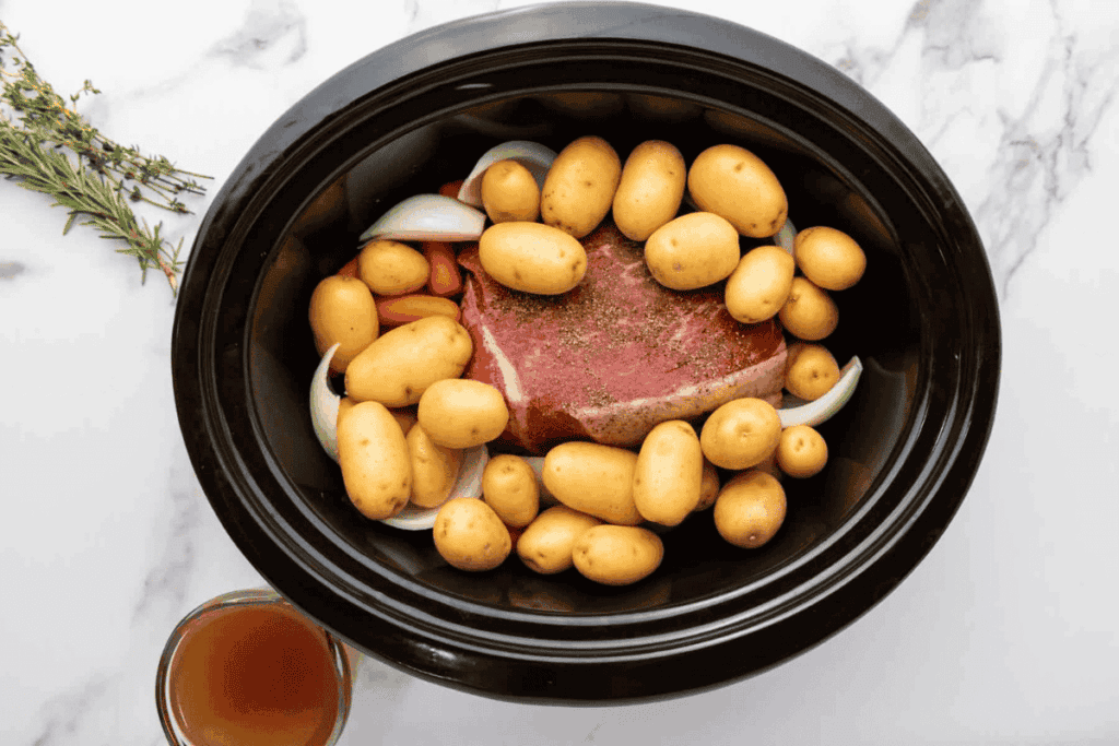 Raw pork roast, baby potatoes, onion wedges, and carrots are arranged in a slow cooker, with a cup of broth on the side and fresh herbs on a marble countertop.