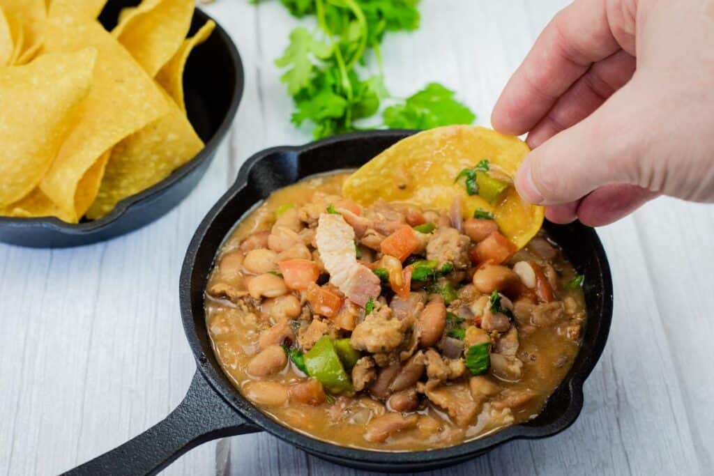 A hand dips a tortilla chip into a small cast-iron skillet filled with Charro Beans With Chorizo, more chips and cilantro are in the background.