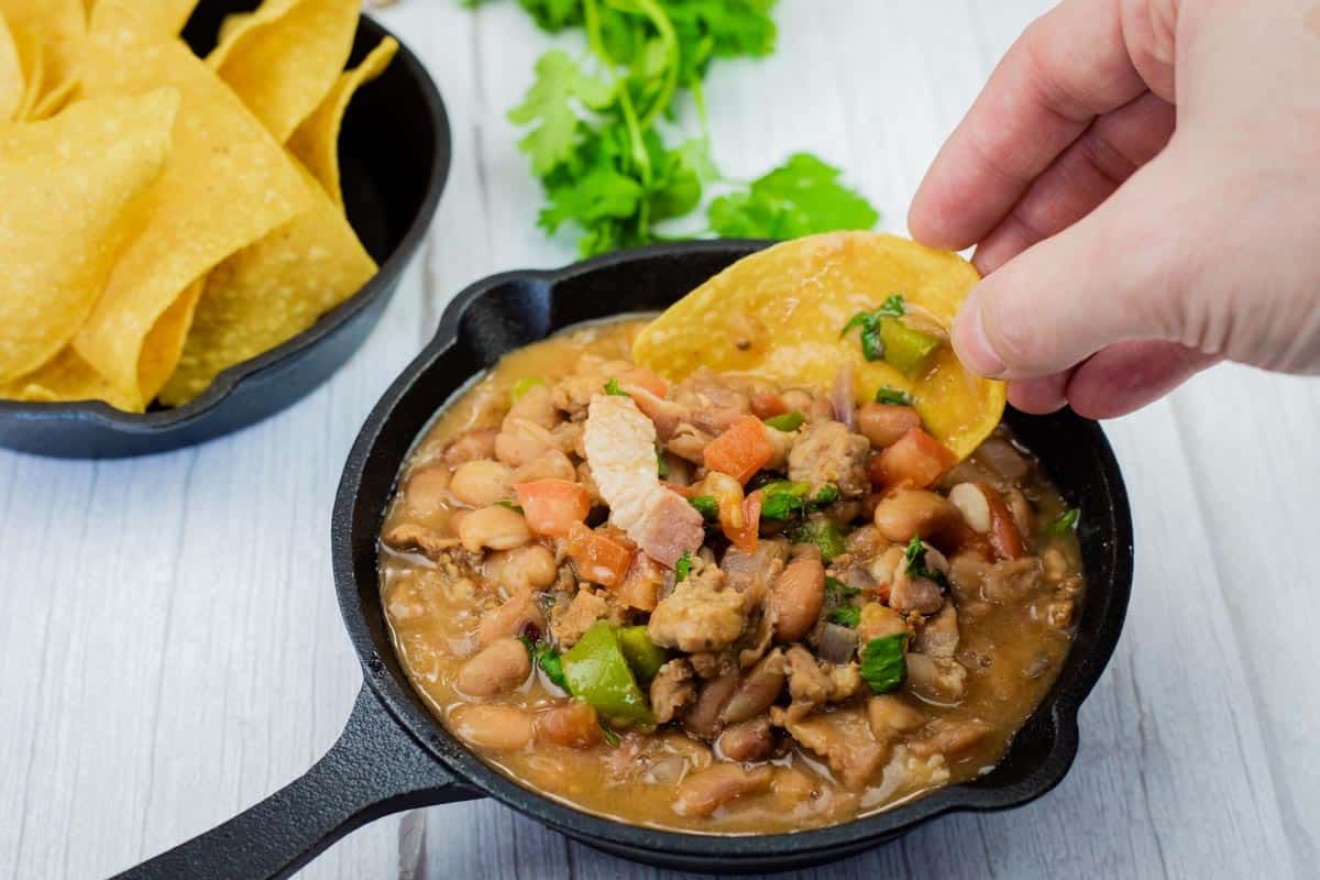 A hand dips a tortilla chip into a small cast-iron skillet filled with Charro Beans With Chorizo, more chips and cilantro are in the background.
