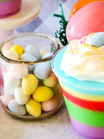 A glass jar of pastel candy eggs, a cup of colorful layered dessert topped with whipped cream and a candy egg, and plastic Easter eggs on a light surface.