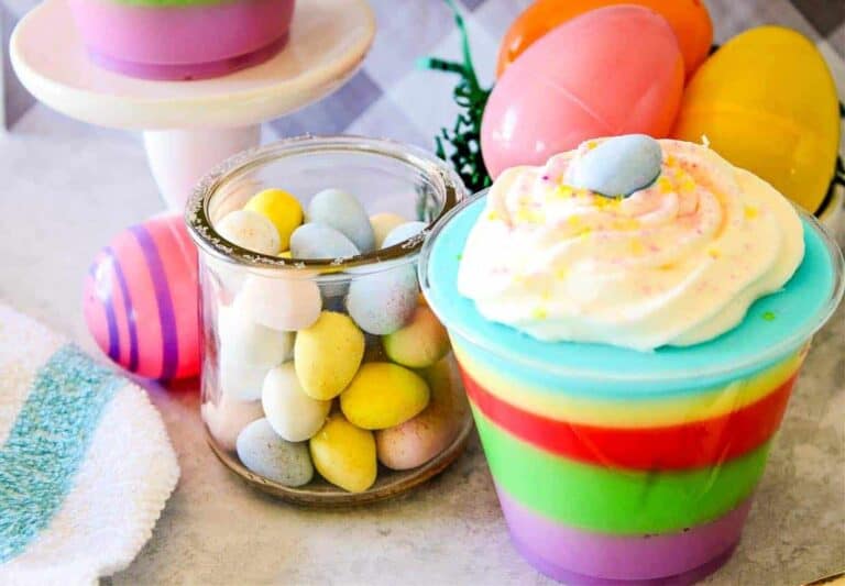 A glass jar of pastel candy eggs, a cup of colorful layered dessert topped with whipped cream and a candy egg, and plastic Easter eggs on a light surface.