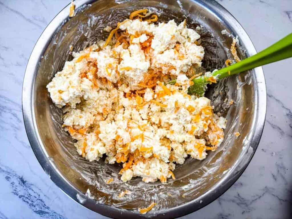 A metal mixing bowl filled with a mixture of shredded cheese, cream cheese, and mayonnaise being stirred with a green spatula.