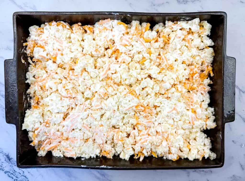 A baking pan filled with an uncooked mixture of shredded cheese, potatoes, and a creamy sauce, spread evenly before baking.