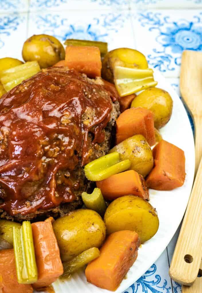 Meatloaf topped with sauce, surrounded by cooked potatoes, carrots, and celery on a white plate.