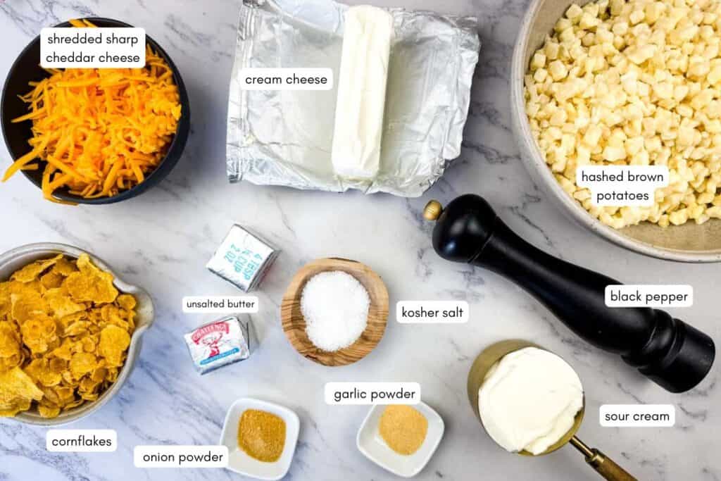 Ingredients for a casserole are arranged on a countertop: shredded cheddar, cream cheese, hashbrowns, butter, salt, black pepper, cornflakes, garlic powder, onion powder, and sour cream.