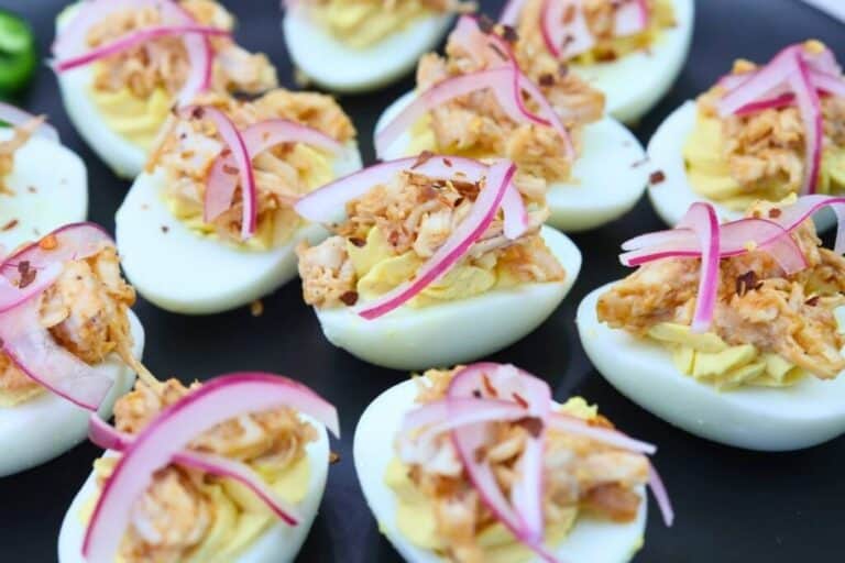 Halved boiled eggs topped with a creamy filling, shredded meat, and thin slices of red onion, arranged on a dark tray.