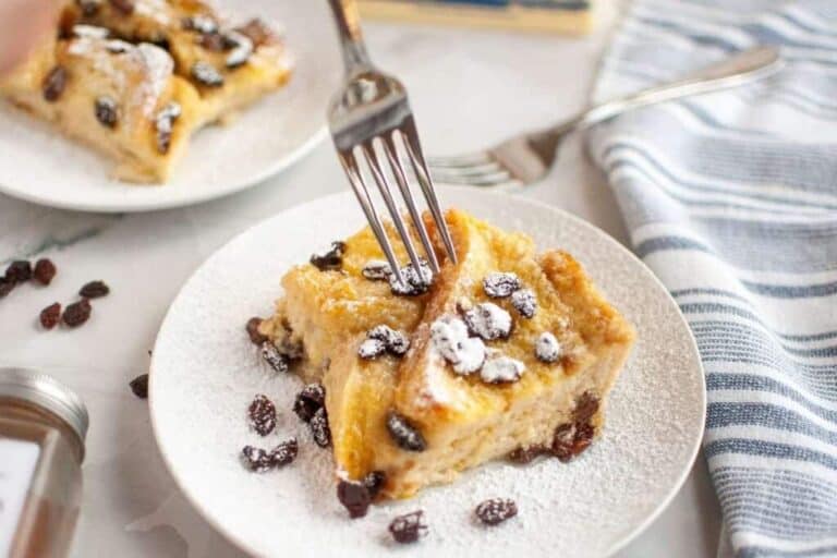 A fork cutting into two slices of bread pudding with raisins, topped with powdered sugar on a white plate.