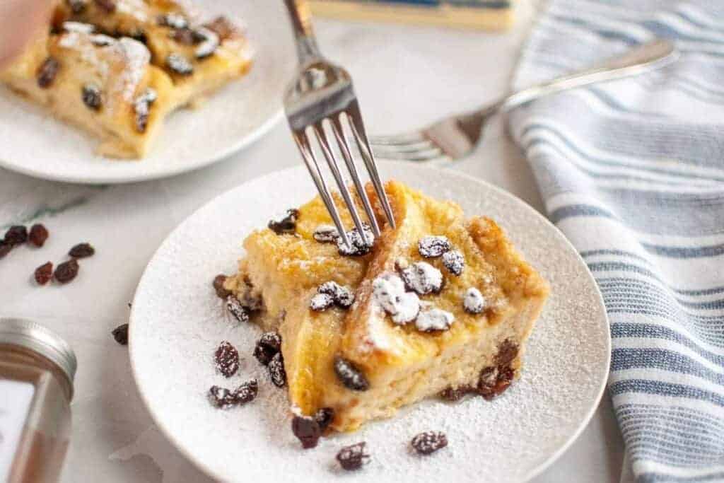 A fork cutting into a slice of bread pudding with raisins, dusted with powdered sugar, served on a white plate.