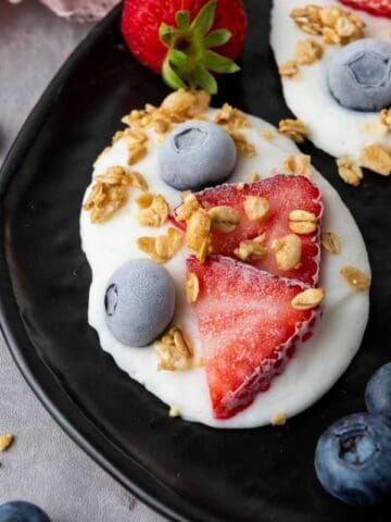 Three Frozen Vanilla Yogurt Bites topped with sliced strawberries, blueberries, and granola are arranged on a black plate, with extra berries and granola scattered around on the gray surface.