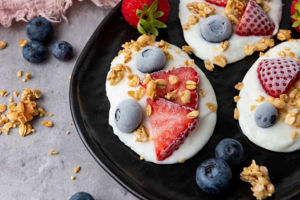 Three Frozen Vanilla Yogurt Bites topped with sliced strawberries, blueberries, and granola are arranged on a black plate, with extra berries and granola scattered around on the gray surface.