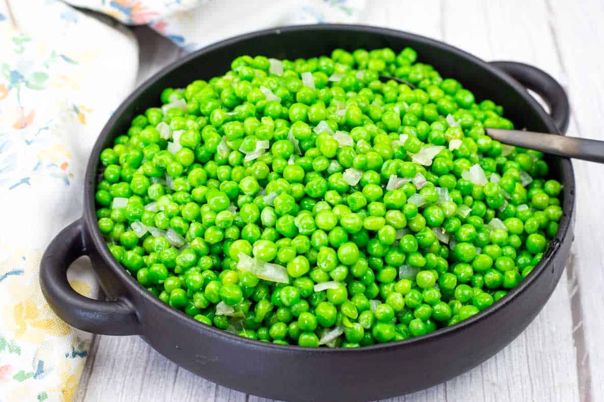 A black bowl filled with cooked green peas and chopped onions sits on a light wooden surface with a spoon inside.