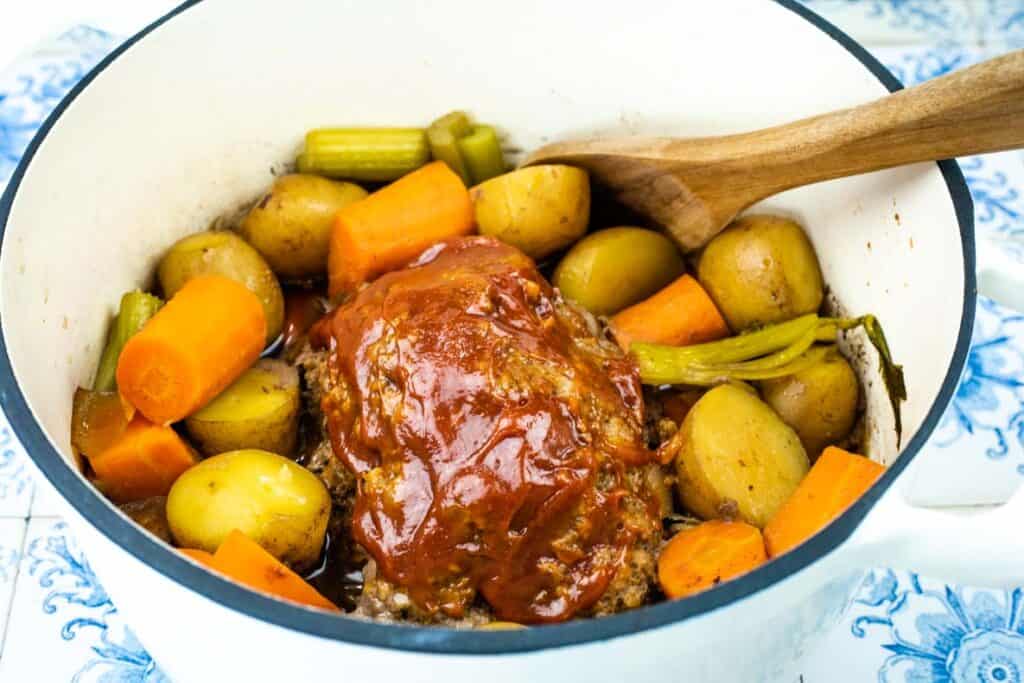 A white Dutch oven filled with glazed meatloaf, whole baby potatoes, carrot chunks, and celery sticks, with a wooden spoon resting on the edge.