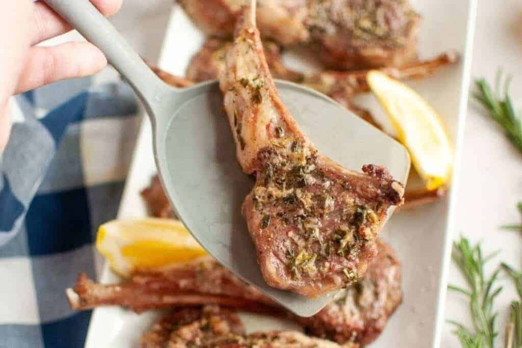 A cooked lamb chop with herbs is lifted by a spatula above a plate with more lamb chops and lemon wedges.