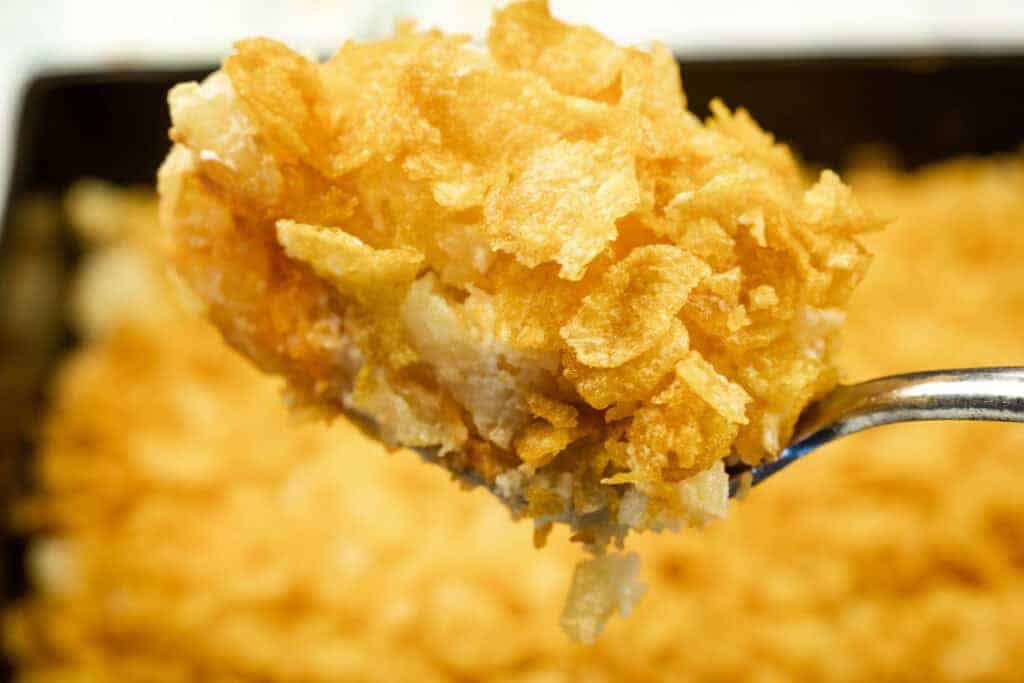 A close-up of a spoonful of Funeral potatoes with Cream Cheese, topped with crushed cornflakes, with the rest of the casserole in the background.