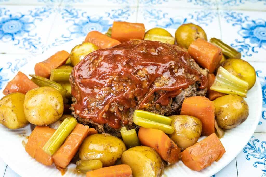 A plate of meatloaf topped with ketchup, surrounded by cooked potatoes, carrots, and celery on a white plate.