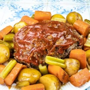 A plate of meatloaf topped with ketchup, surrounded by cooked potatoes, carrots, and celery on a white plate.