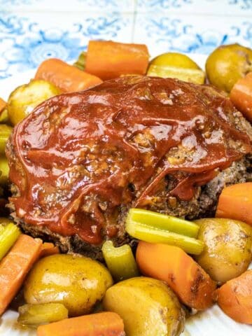 A plate of meatloaf topped with ketchup, surrounded by cooked potatoes, carrots, and celery on a white plate.