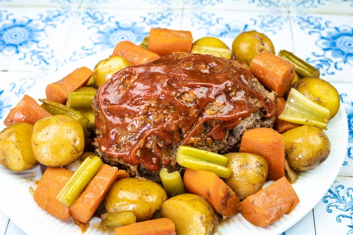 A plate of meatloaf topped with tomato sauce, surrounded by cooked baby potatoes, carrots, and celery on a patterned tile background.