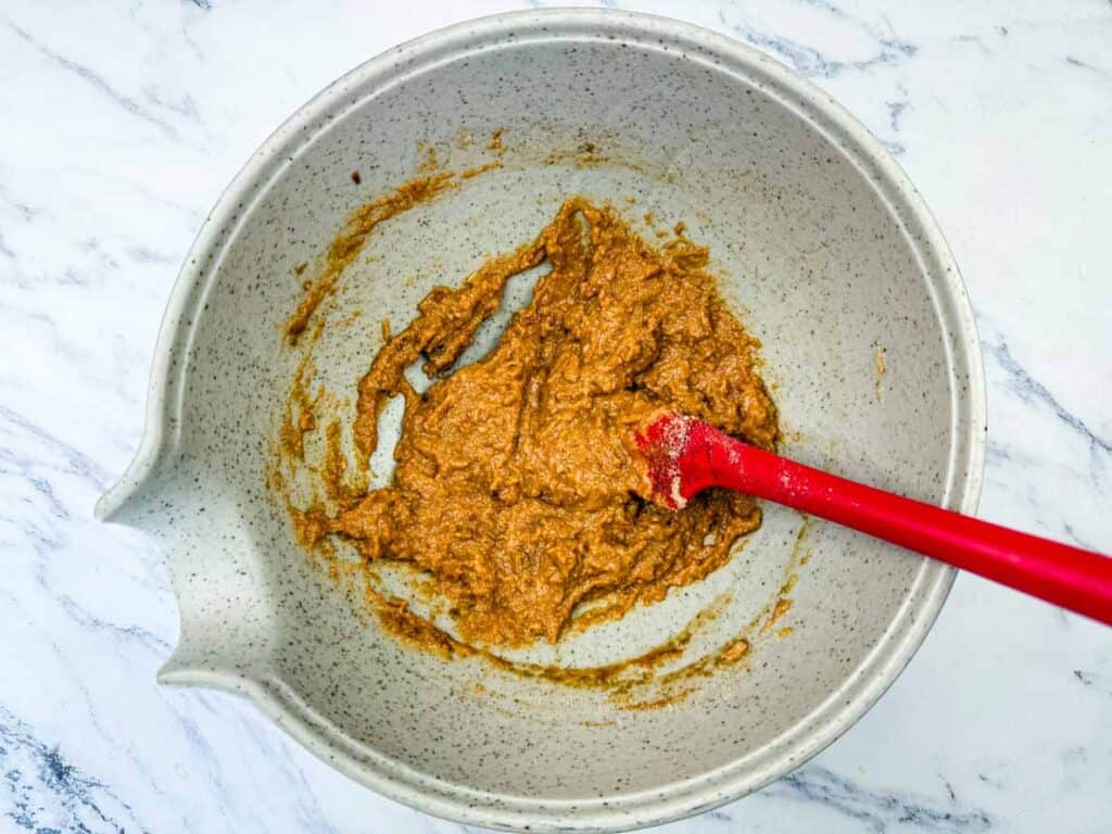 A red spatula rests in a white mixing bowl containing thick brown batter on a marble surface.