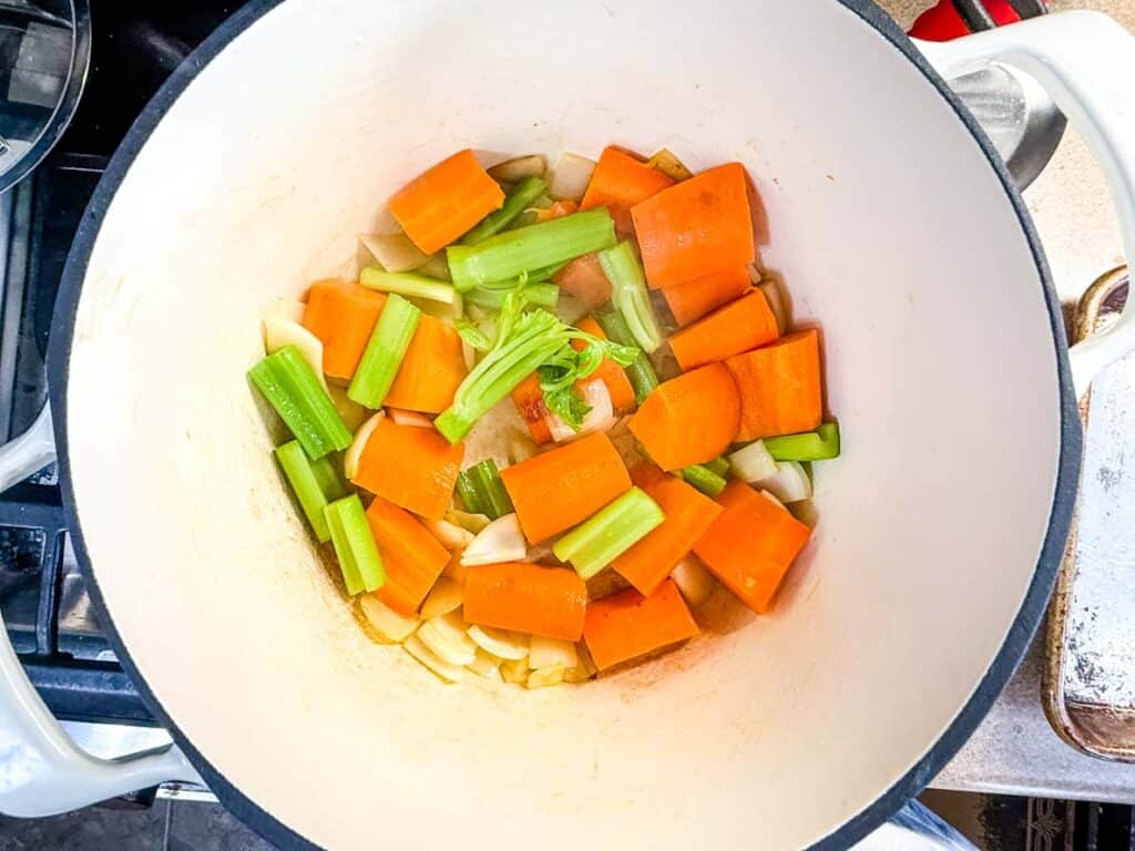 Chopped carrots, celery, and onions in a white pot on a stovetop, beginning to be sautéed for cooking.