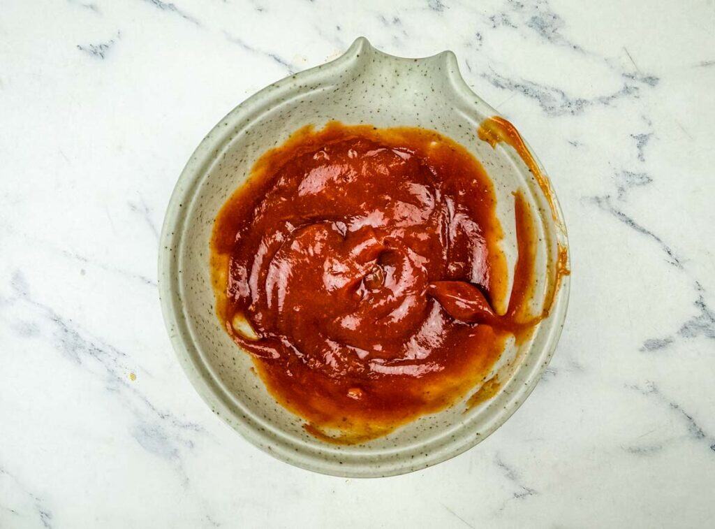 A bowl filled with thick reddish-brown barbecue sauce sits on a white marble surface.