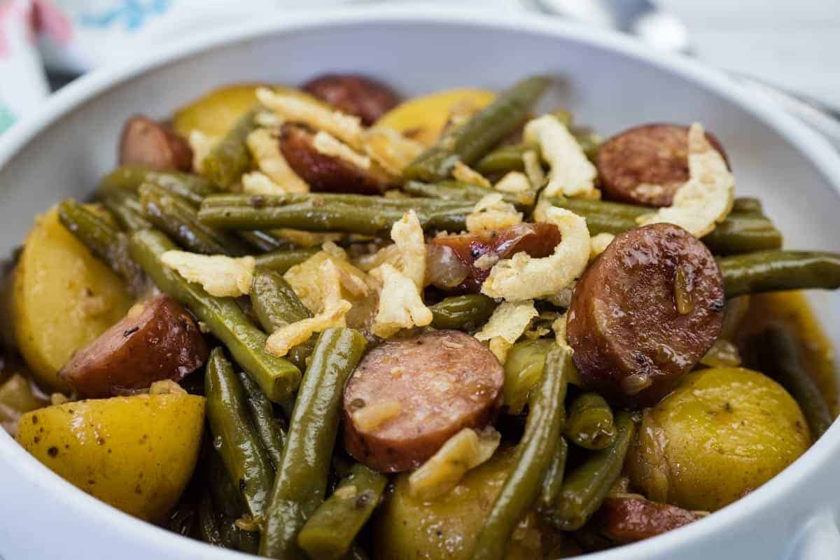 A bowl of Swamp Potatoes with cooked green beans, sliced sausage, and halved potatoes, topped with crispy fried onions.