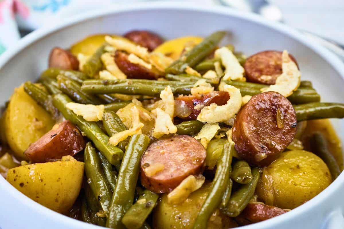 A bowl of Swamp Potatoes with cooked green beans, sliced sausage, and halved potatoes, topped with crispy fried onions.