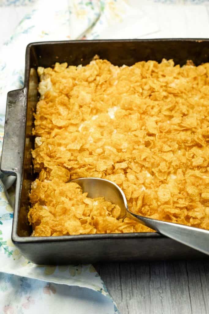 A metal baking pan filled with a casserole topped with golden cornflakes, with a large serving spoon resting in one corner.