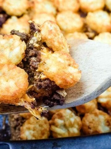 A wooden spatula lifts a serving of tater tot casserole with ground beef and melted cheese from a baking dish.