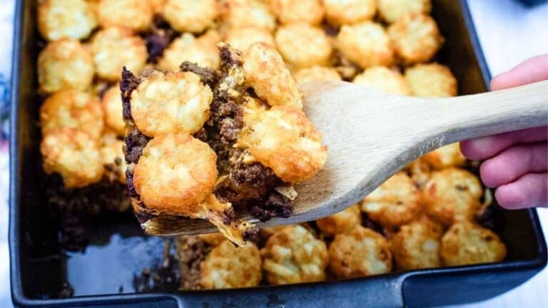 A wooden spatula lifts a serving of tater tot casserole with ground beef and melted cheese from a baking dish.