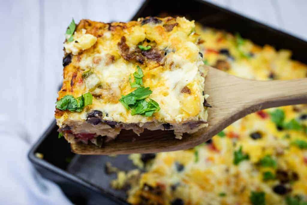 A wooden spatula holds up a square slice of baked casserole topped with melted cheese and chopped herbs, with more casserole in the background.